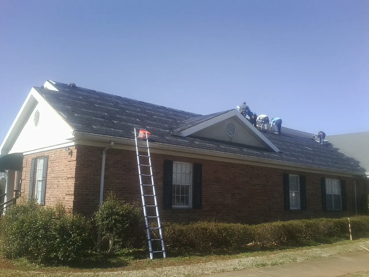 Crew tearing shingles off a brick ranch roof with a ladder set — Metal Roof Repair work by our crew in Blairsburg
