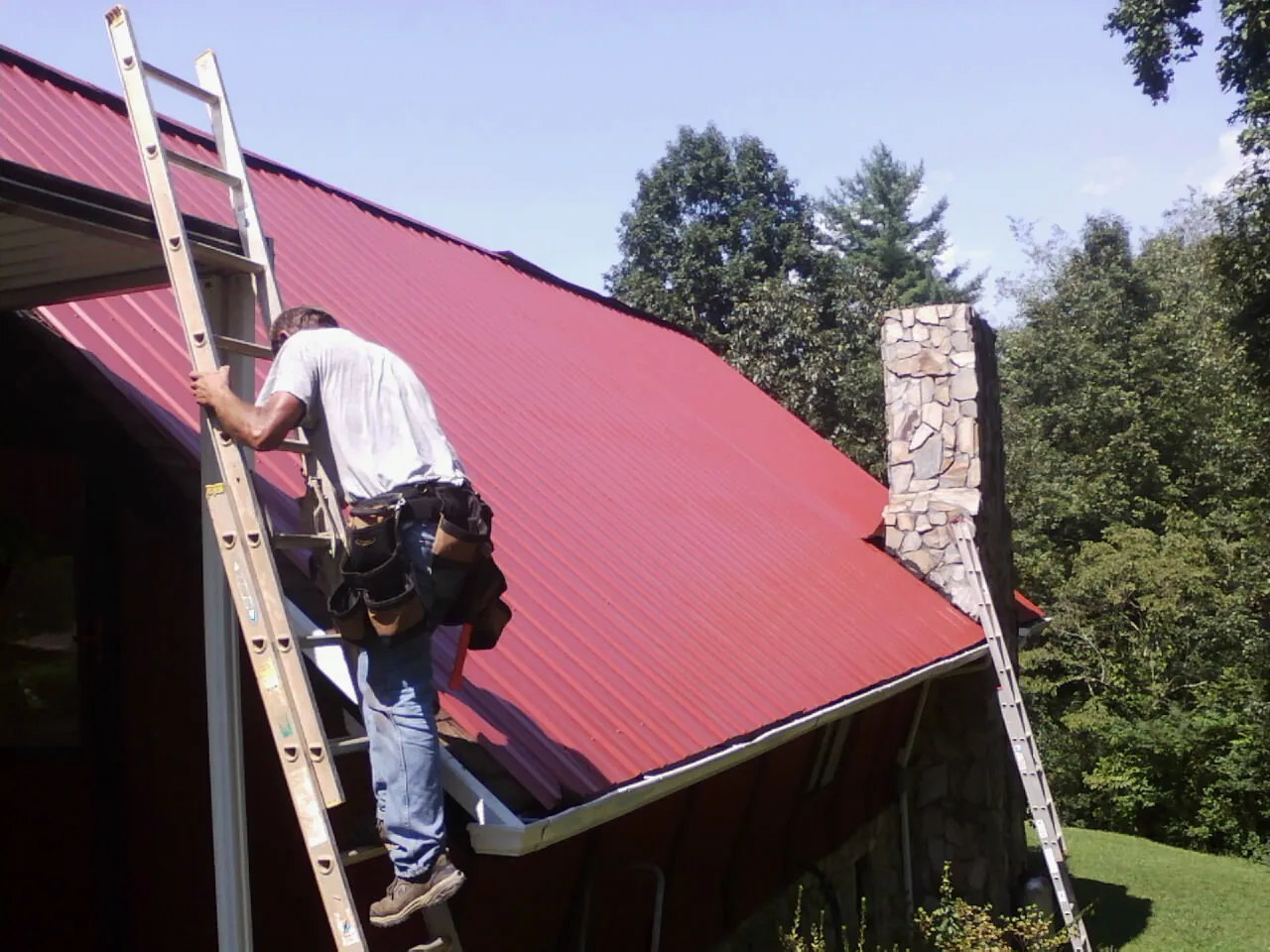Roofer on a ladder beside a red metal roof and stone chimney — Soffit & Fascia Repair by Roofing Brothers in Blairsburg